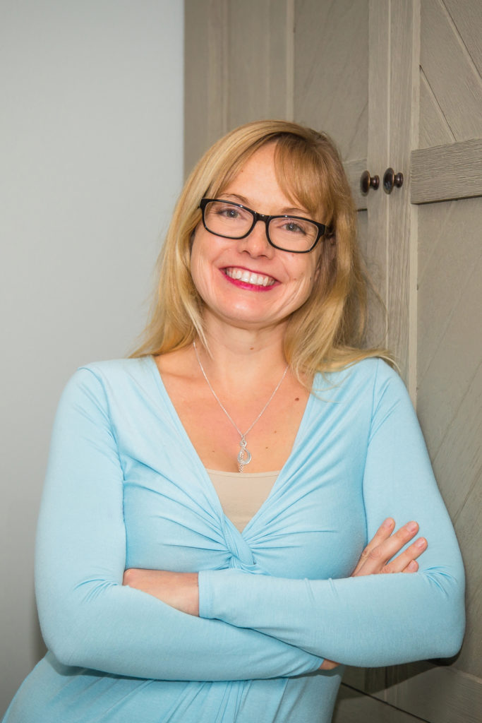 Blonde woman (me!) with  glasses, red lipstick, and blue shirt leaning against a door with her arms crossed.